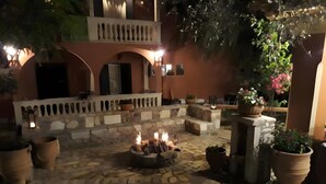 An outdoor patio area with stone flooring, a fire pit, potted plants, and a balcony with a railing. - Comfy Stay Gouvia – Kitchenette 4 (Kontokali)