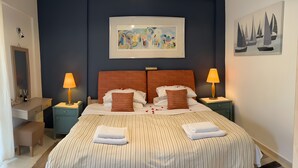 A double bed with striped bedding, two nightstands with lamps, a framed abstract painting above the bed, and a sailboat painting on the wall. - Comfy Stay Gouvia – Kitchenette 4 (Kontokali)