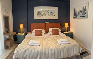 Una cama doble con ropa de cama a rayas, dos mesitas de noche con lámparas, un cuadro abstracto enmarcado sobre la cama y un cuadro de un velero en la pared.