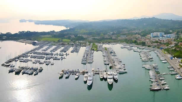 An aerial view of a marina with numerous boats docked, surrounded by a coastal town with buildings and greenery. - Comfy Stay Gouvia – Kitchenette 4 (Kontokali)