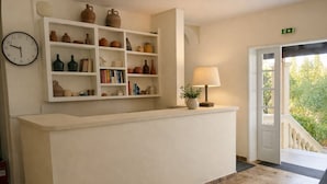 A reception desk with a clock, shelves with pottery and books, a lamp, and a door leading outside. - Marina Balcony – Kitchenette 8 (Kontokali)