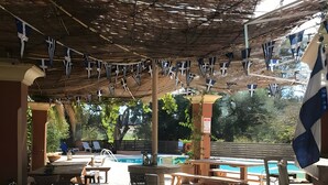 An outdoor pool area with stone flooring, lounge chairs, and a pergola with flags. - Marina Balcony – Kitchenette 8 (Kontokali)