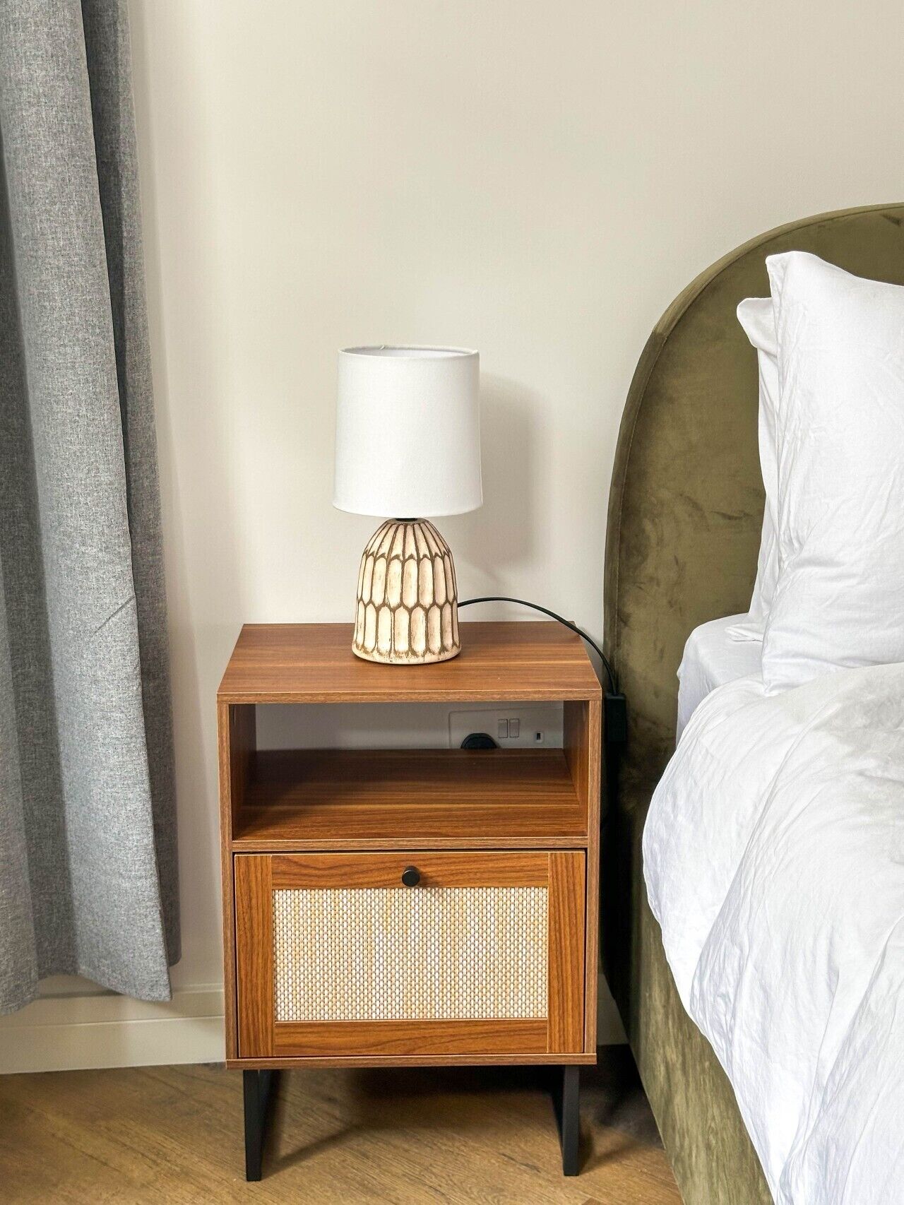 A wooden bedside table with a lamp, a drawer, and a bed with white bedding.