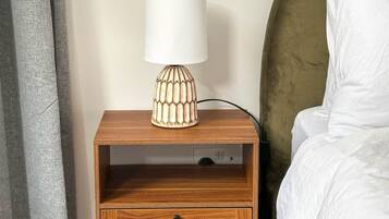 A wooden bedside table with a lamp, a drawer, and a bed with white bedding.