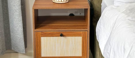 A wooden bedside table with a lamp, a drawer, and a bed with white bedding.