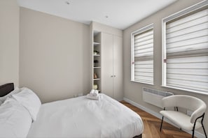 A bedroom with a bed, a chair, a wardrobe, and a window with blinds. - Studio, West Kensington + Hammersmith (Greater London)