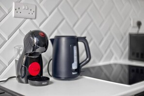 Eine Kaffeemaschine, ein Wasserkocher und ein Glas auf einer Arbeitsplatte mit weiß gefliestem Rücksprühschutz.