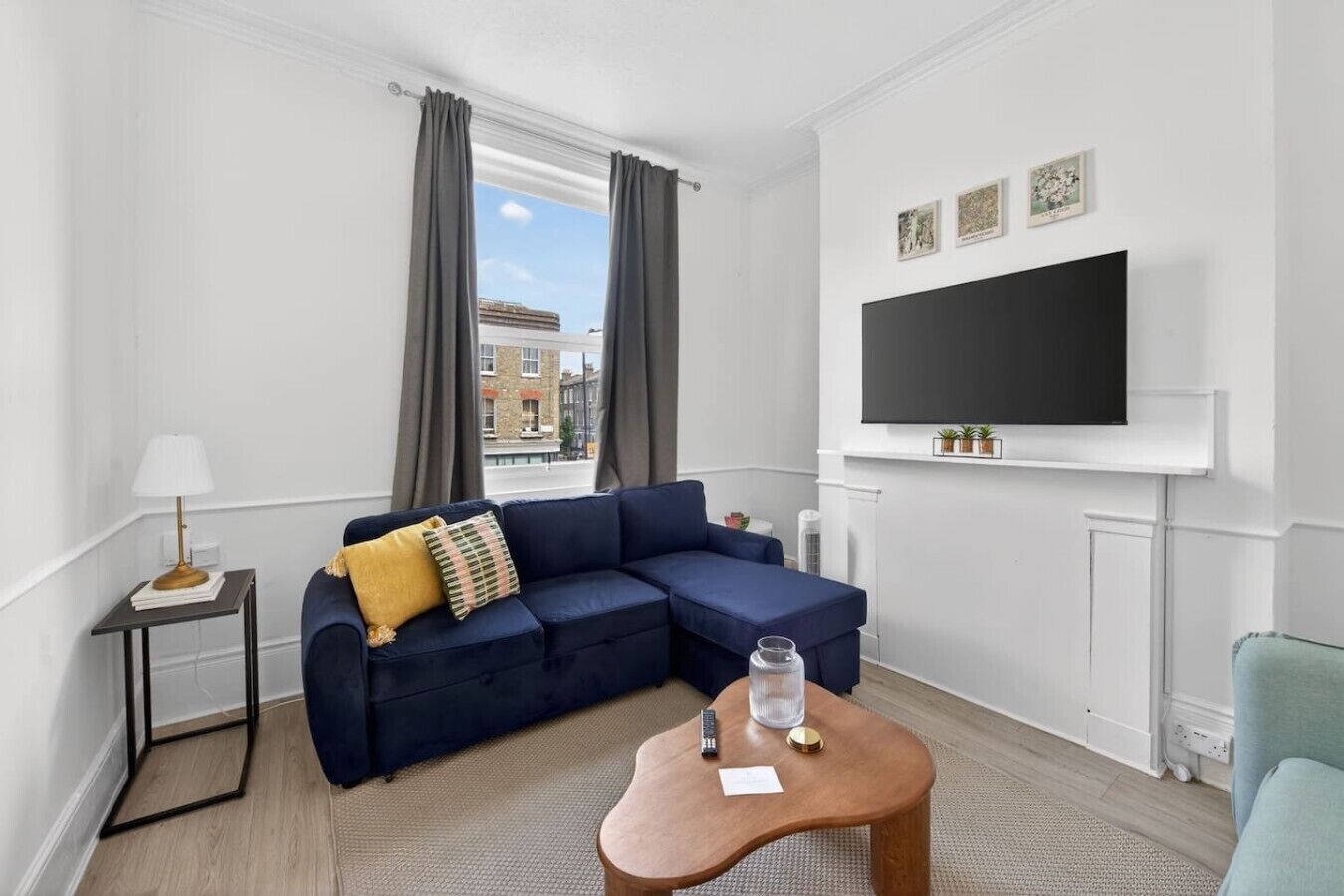 A modern living room with a sectional sofa, a small side table, a mounted TV, and a view of buildings outside the window.
