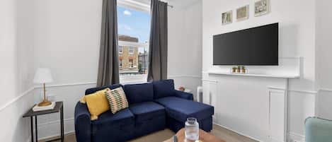 A modern living room with a sectional sofa, a small side table, a mounted TV, and a view of buildings outside the window.