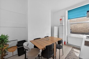 A small, modern room with a wooden table and four chairs, a radiator, a window with a view of a brick building, and a fire extinguisher on the wall. - Finsbury Park | Stylish 1BR | Near Tube | Wifi (Greater London)
