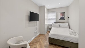 A bedroom with a bed, a TV mounted on the wall, a chair, and a window with blinds. - Studio, West Kensington + Hammersmith (Greater London)