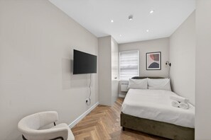 Quarto com cama, televisão na parede, cadeira e janela com persianas.