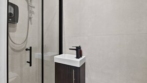 A modern bathroom with a glass shower enclosure, a white sink with a dark cabinet, and a control panel on the wall. - Modern Studio in Barons Court | Near Kensington & Hammersmith (Greater London)