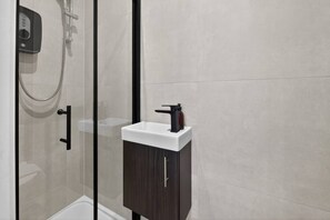 Casa de banho moderna com cabine de duche em vidro, lavatório branco com armário escuro e painel de controlo na parede.