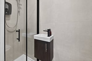 A modern bathroom with a glass shower enclosure, a white sink with a dark cabinet, and a control panel on the wall. - Studio, West Kensington + Hammersmith (Greater London)