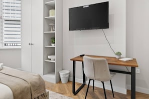 A bedroom with a bed, a desk with a chair, a TV mounted on the wall, a window with blinds, and a white wardrobe. - Studio, West Kensington + Hammersmith (Greater London)