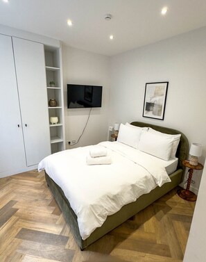 A neatly made bed with white linens and a folded towel, a television mounted on the wall, a framed picture, and a bedside table with a lamp. - Modern Studio in Barons Court | Near Kensington & Hammersmith (Greater London)