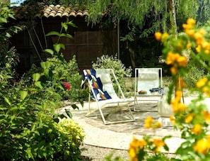 Un jardín con dos sillones de descanso, una estructura con techo de tejas y abundante vegetación.