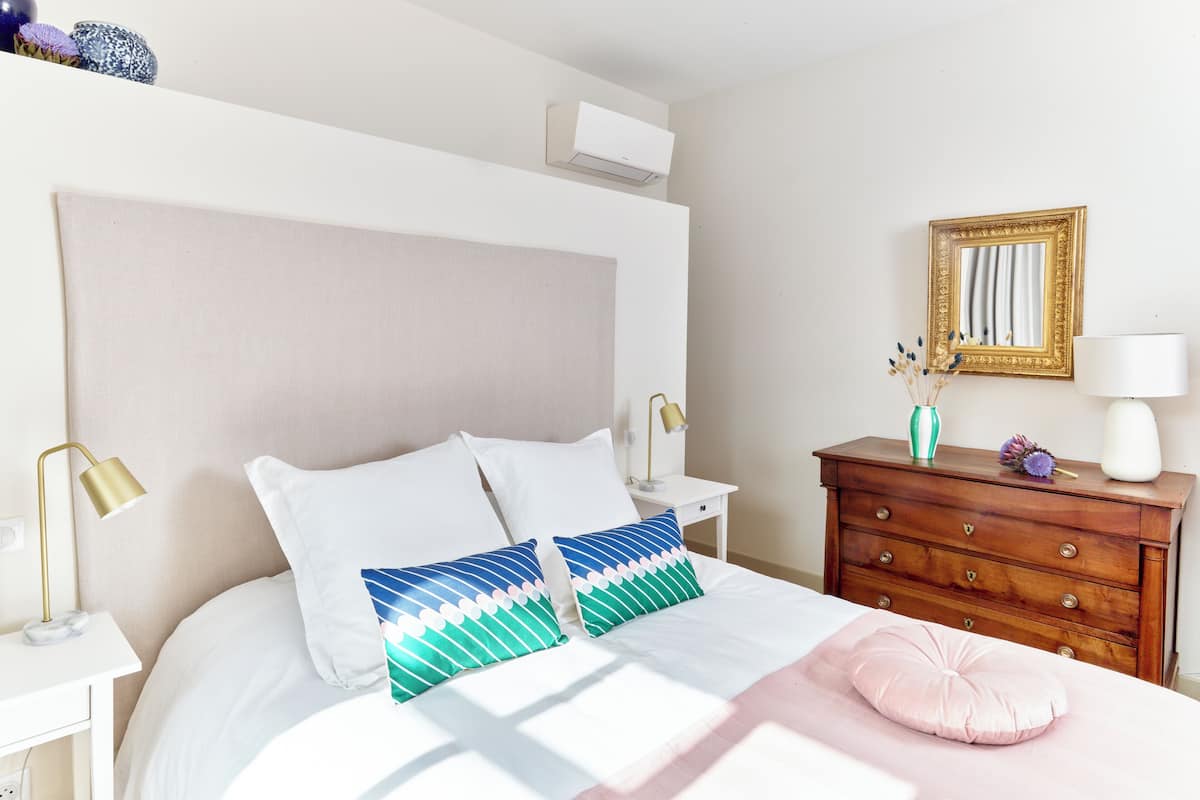 A bedroom with a bed, bedside tables, a dresser, a mirror, and an air conditioning unit.