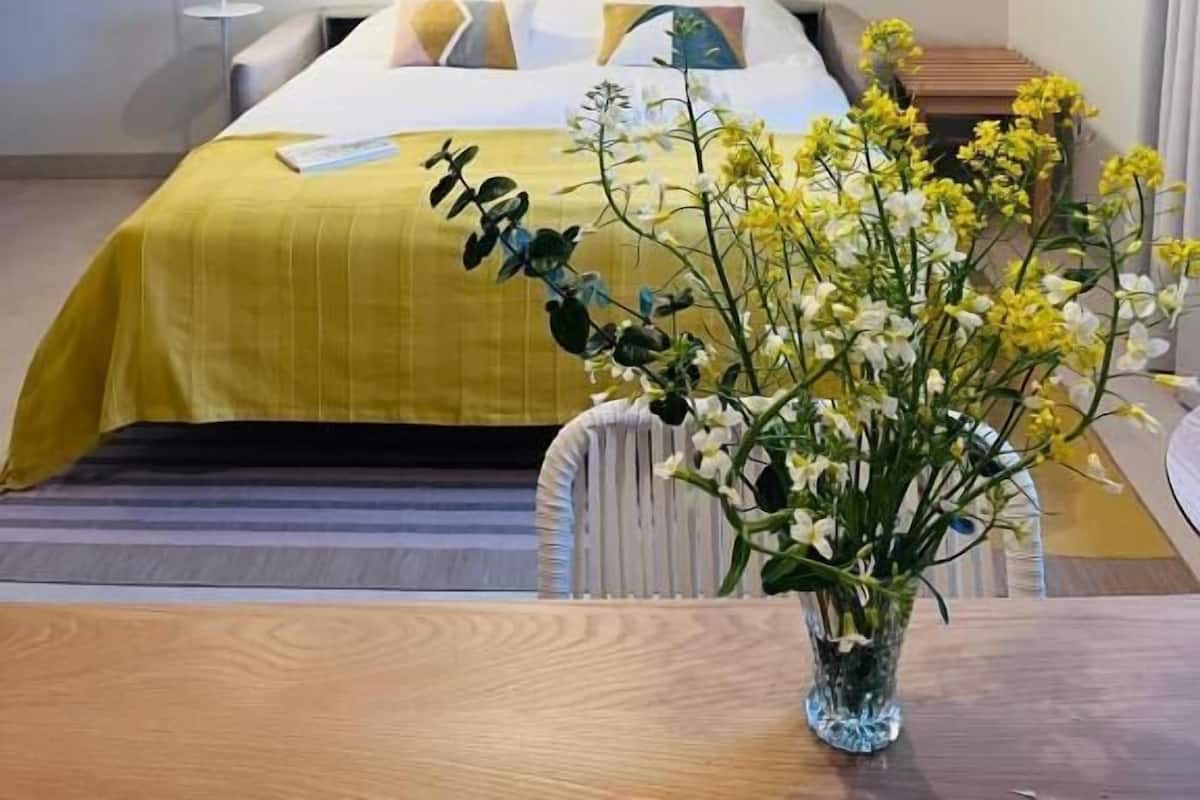 A hotel room with a bed, a bedside table, a vase of flowers, a hanging light fixture, and framed pictures on the wall.