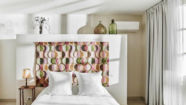 A bedroom with a patterned headboard, a bedside table with a lamp, and a window with curtains. - Château de la Piscine - Appartements dans le Mas au coeur d'un grand parc arboré (Montpellier)