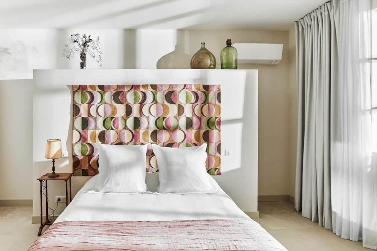 A bedroom with a patterned headboard, a bedside table with a lamp, and a window with curtains.
