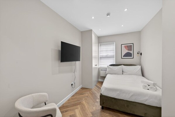 A bedroom with a bed, a TV mounted on the wall, a chair, and a window with blinds. - Studio, West Kensington + Hammersmith (Greater London)