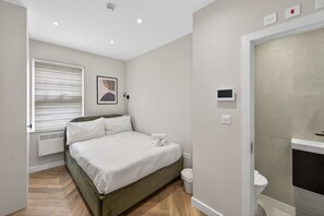 A bedroom with a bed, a window with blinds, a wall-mounted TV, and a toilet. - Studio, West Kensington + Hammersmith (Greater London)