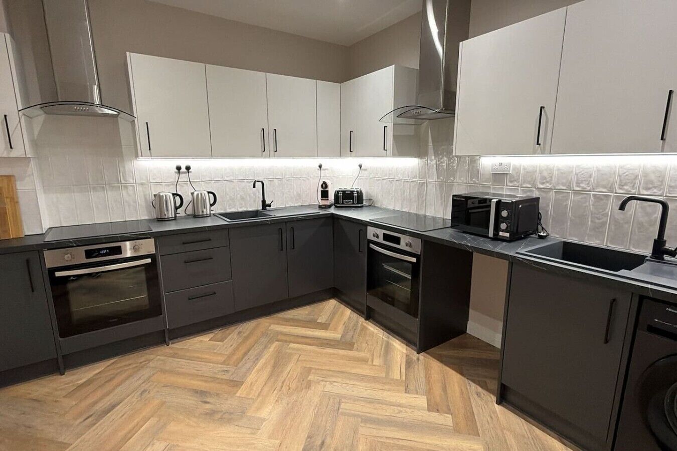 A modern kitchen with dark cabinetry, stainless steel appliances, and a herringbone wood floor.
