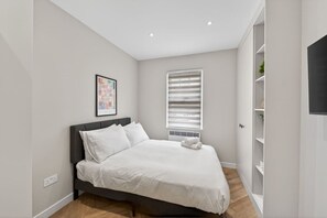 A bedroom with a bed, a window with blinds, a wall-mounted TV, and a shelf with items. - Studio, West Kensington + Hammersmith (Greater London)