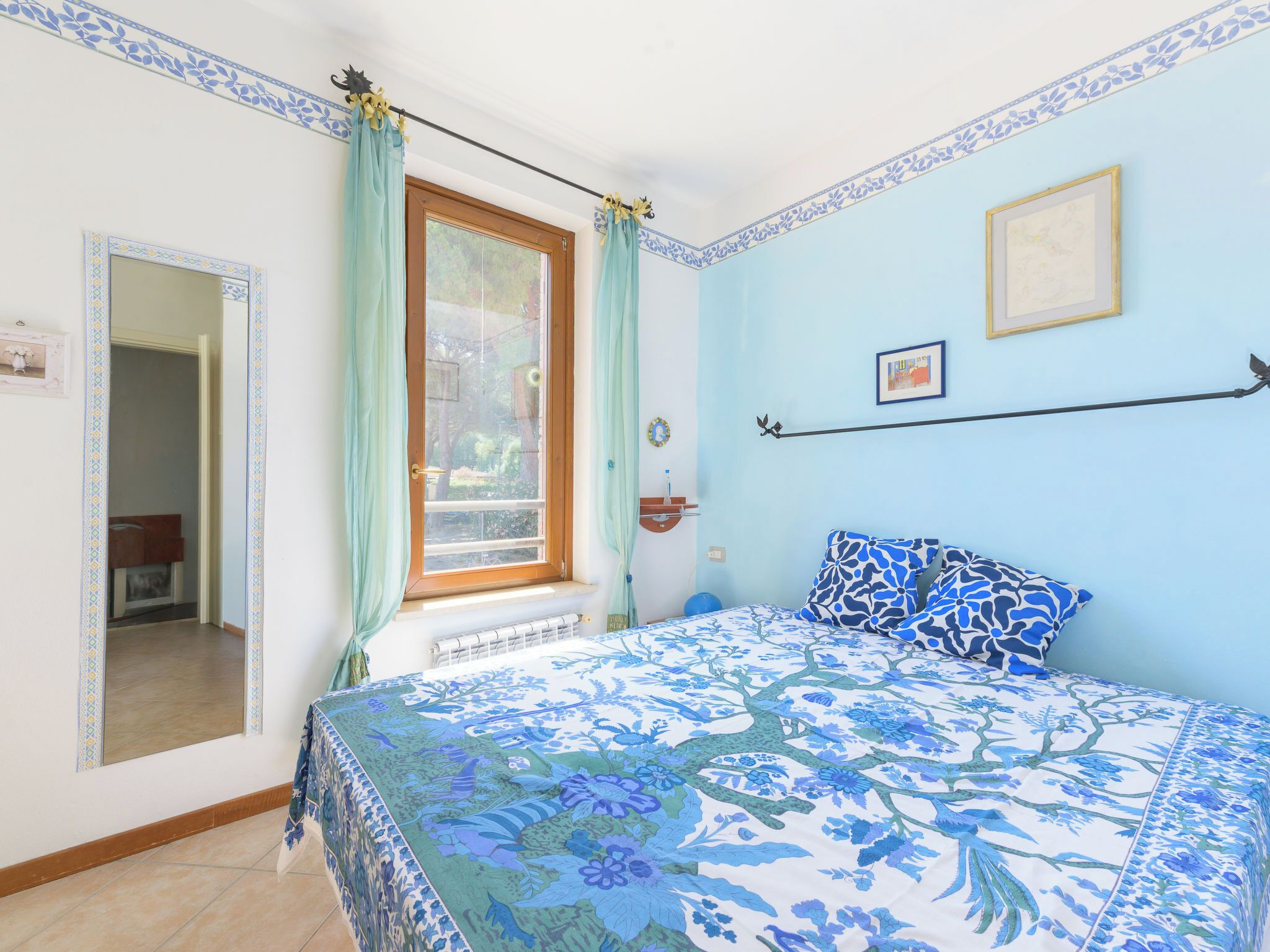 Un dormitorio con cama, ventana con cortinas, radiador, un cuadro en la pared y una puerta que da a otra habitación.