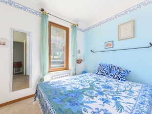 Een slaapkamer met een bed, een raam met gordijnen, een radiator, een foto aan de muur en een deur naar een andere kamer.
