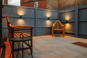 Ein Terrassenbereich im Freien mit einer hölzernen Bank und einem kleinen Tisch, begrenzt von einem Holzzaun mit Wandleuchten.