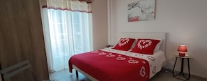Ein Schlafzimmer mit einem Bett, einem Nachttisch, einer Lampe, einem Bild an der Wand und einem Fenster mit durchsichtigen Vorhängen.