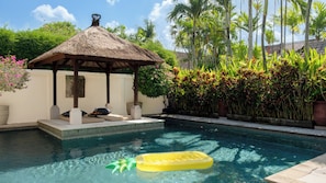 Ein Pool mit einer strohgedeckten Struktur, Liegebereich und tropischen Pflanzen.
