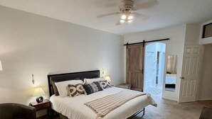 A bedroom with a bed, bedside tables, a chair, a ceiling fan, and a door leading to another room.