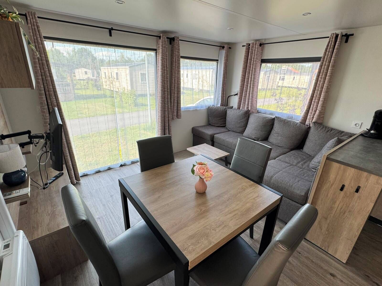 Un intérieur de camping-car moderne comprenant une table à manger, des sièges, un coin cuisine et de grandes fenêtres offrant une vue sur l’extérieur.
