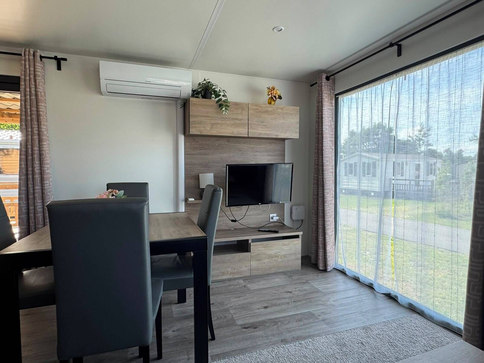A modern living room with a flat-screen TV, wooden cabinets, a dining table, and a large sliding glass door with a view of the outdoors.