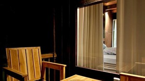 A wooden chair and table with a woven placemat, a large window with curtains, and a glimpse of a bed in the reflection.