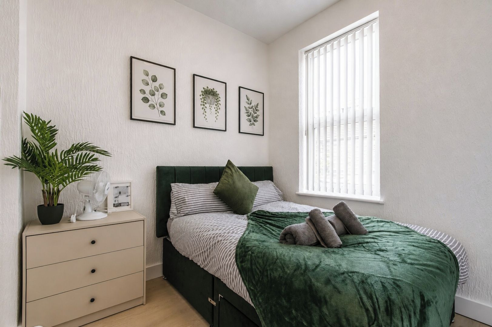 A bedroom with a bed, a bedside table with a plant, a window with blinds, and three framed botanical prints on the wall.