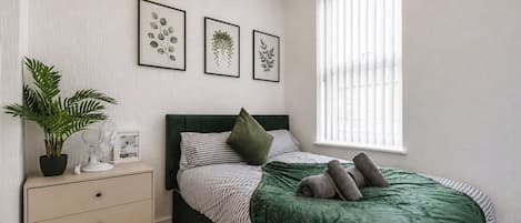 Een slaapkamer met een bed, een nachtkastje met een plant, een raam met jaloezieën en drie ingelijste botanische prints aan de muur.