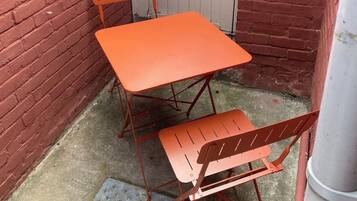 Ein kleiner Außenbereich mit Tisch und zwei Stühlen, einem Metallgitter und einer Backsteinmauer.