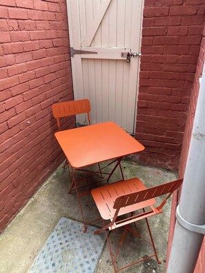 Ein kleiner Außenbereich mit Tisch und zwei Stühlen, einem Metallgitter und einer Backsteinmauer.