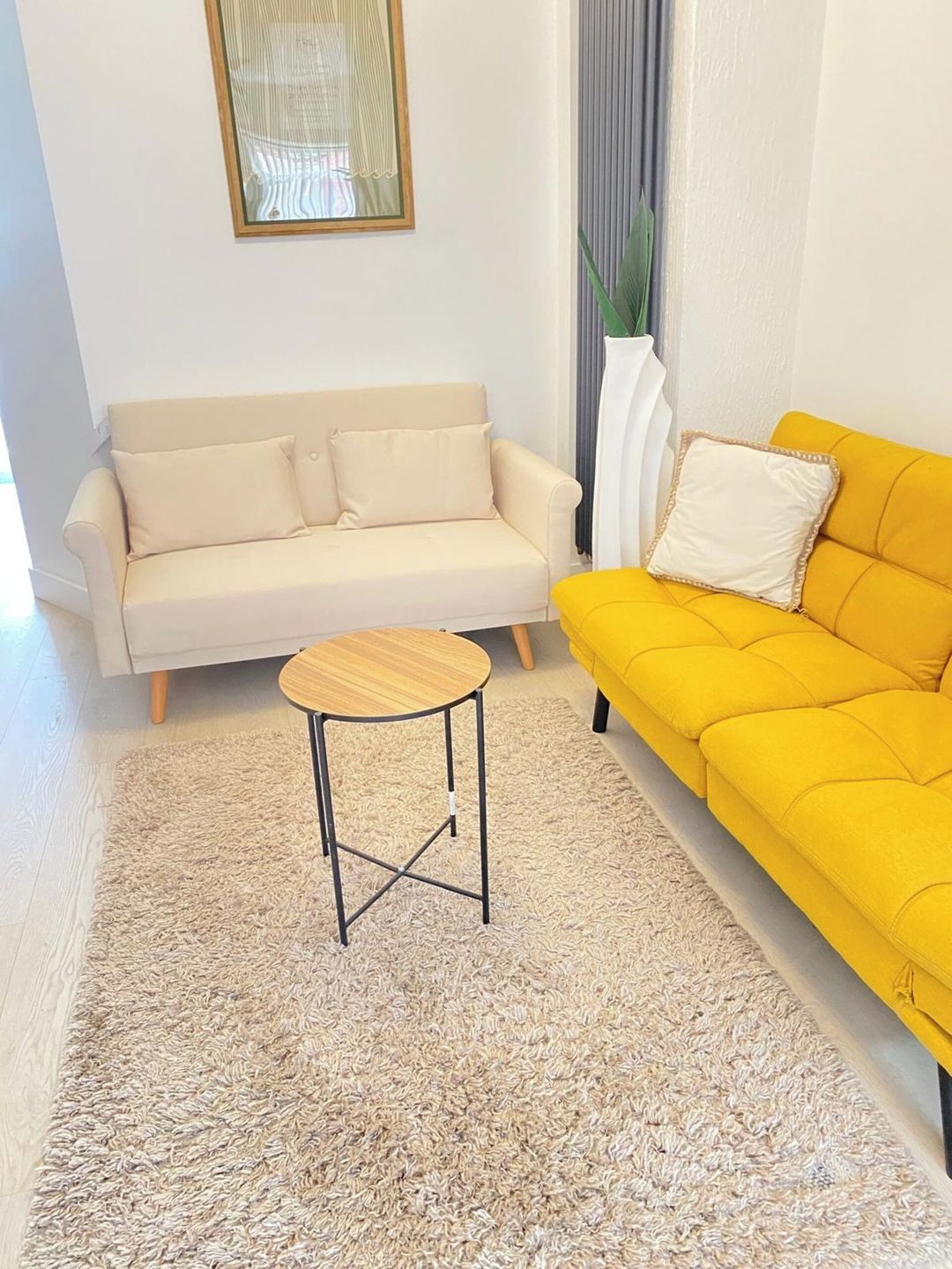 A living room with a beige sofa, a yellow sofa, a small wooden table, and a large potted plant.
