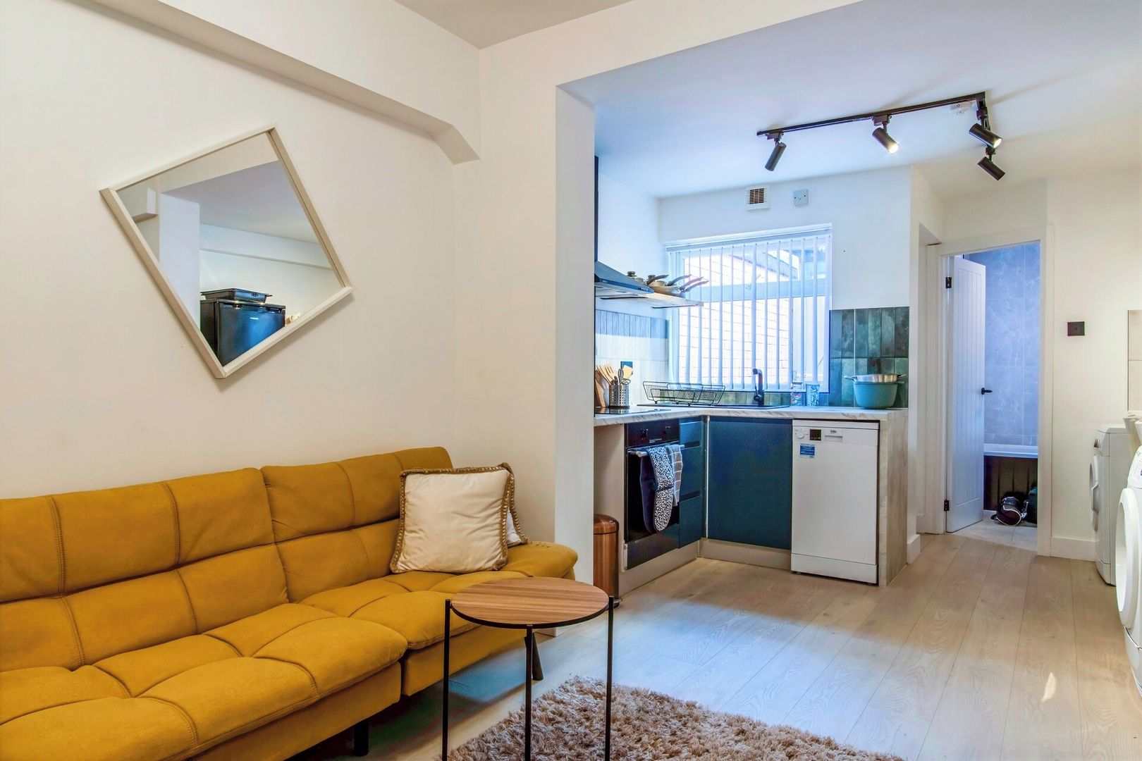 A modern living room with a sofa and a small table, a kitchen with a dishwasher and a window, and a door leading to another room.