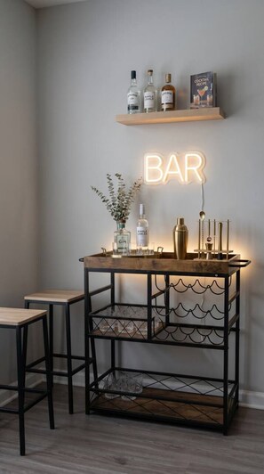 Zona de bar con un letrero de neón, estantes con botellas, una mesa de madera y un carrito de bar.
