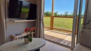 Een kamer met een televisie op een plank, een tafeltje met een bloempot, en uitzicht op een houten terras en groen buiten.