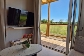 Une pièce avec un téléviseur posé sur une étagère, une petite table sur laquelle se trouve un pot de fleurs, et une vue sur une terrasse en bois et la verdure environnante.