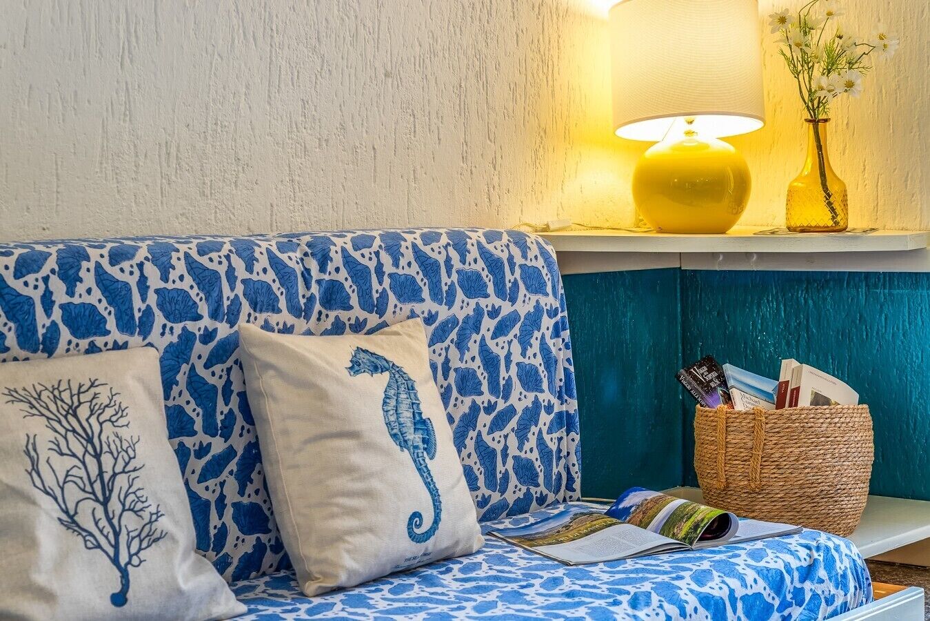 Ein blaues, gemustertes Sofa mit dekorativen Kissen, einer Leselampe und einem Korbwägelchen mit Büchern.