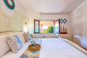 Ein Schlafzimmer mit einem Bett, Kissen, einem Nachttisch mit einem Schiffmodell, einem Fenster mit geöffnetem Klappladen und einem Wanddekor.